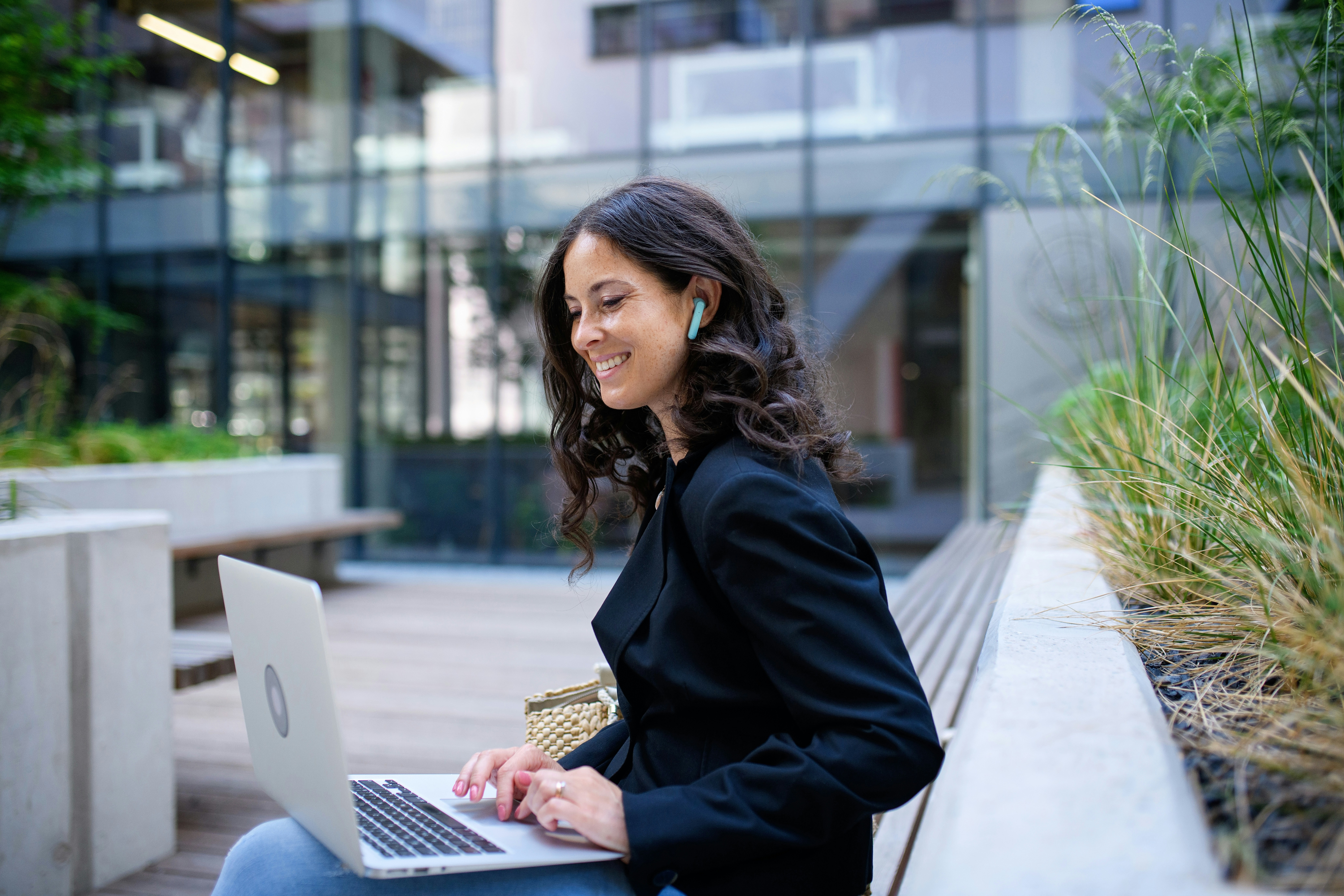 Woman sitting outdoors with a laptop, smiling while working and wearing wireless earbuds.