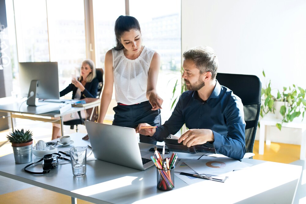 Two colleagues discuss work at a desk with a laptop, while another person works in the background.