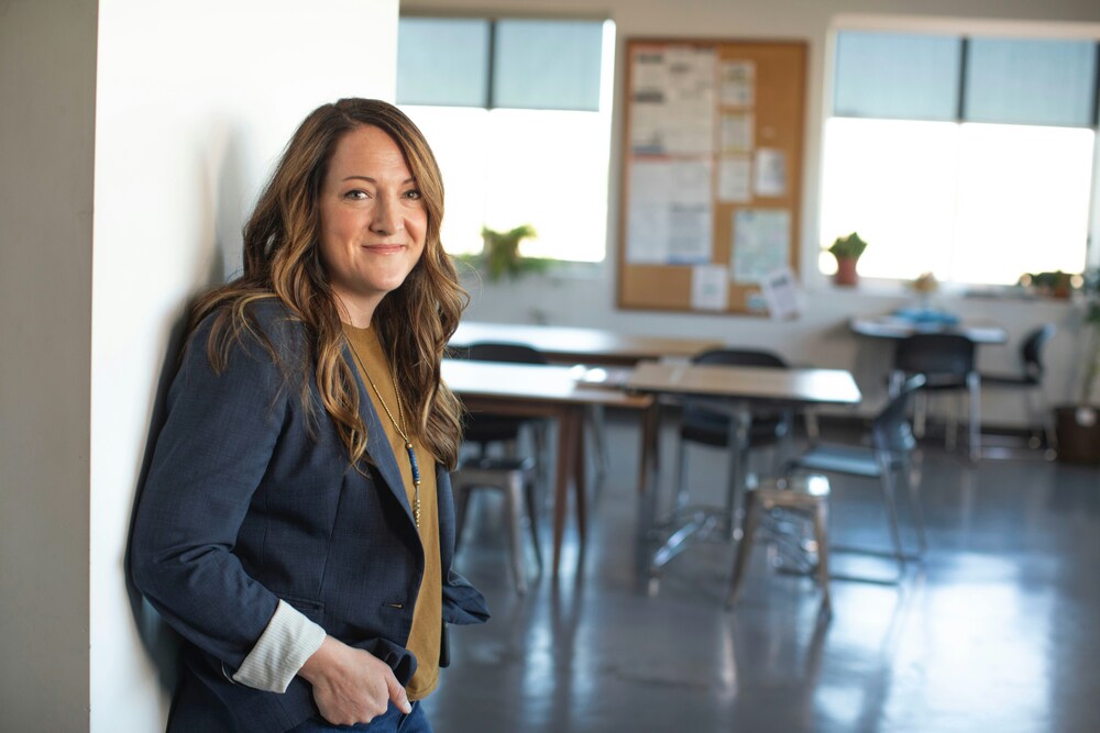 A woman with long hair wearing a blazer leans against a white wall, smiling in a bright office space with tables and chairs in the background.