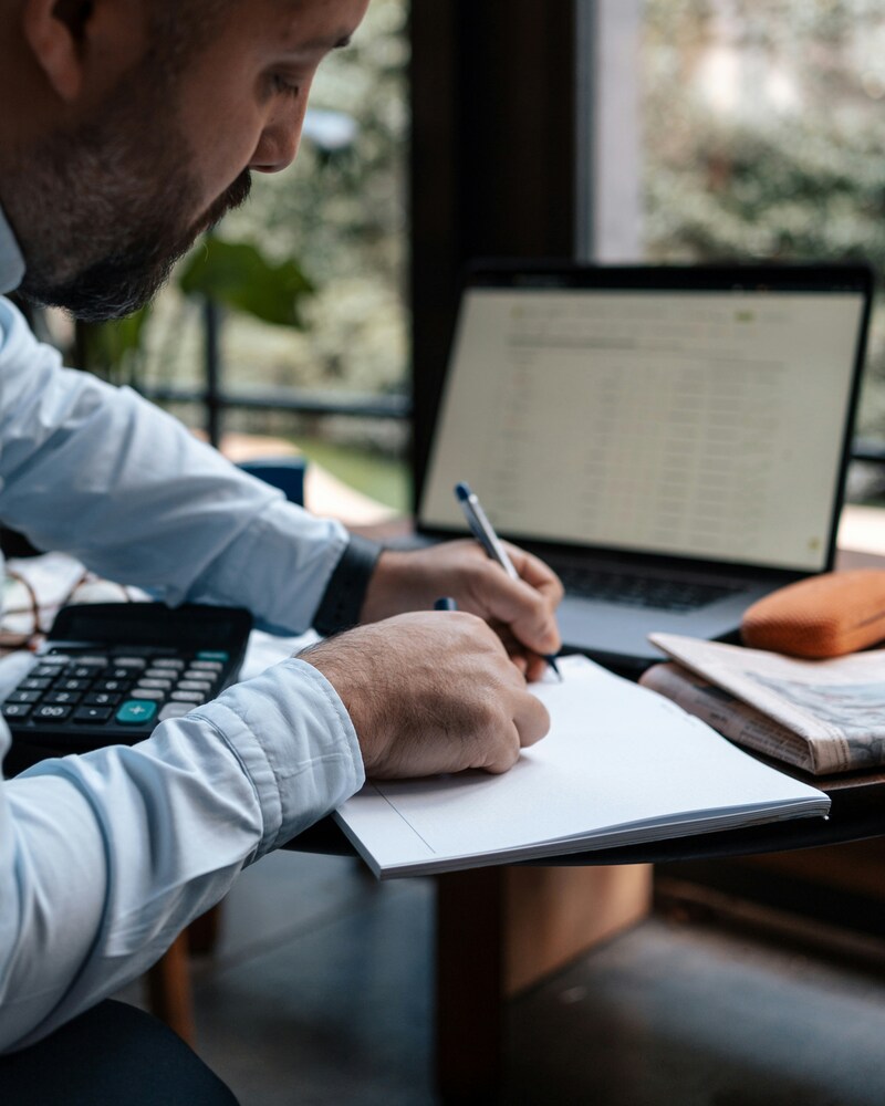 Man writing notes in a notebook at a desk with a laptop and calculator.