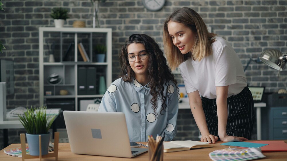 Two women collaborating at a desk, looking at a laptop screen and discussing work in a modern office.